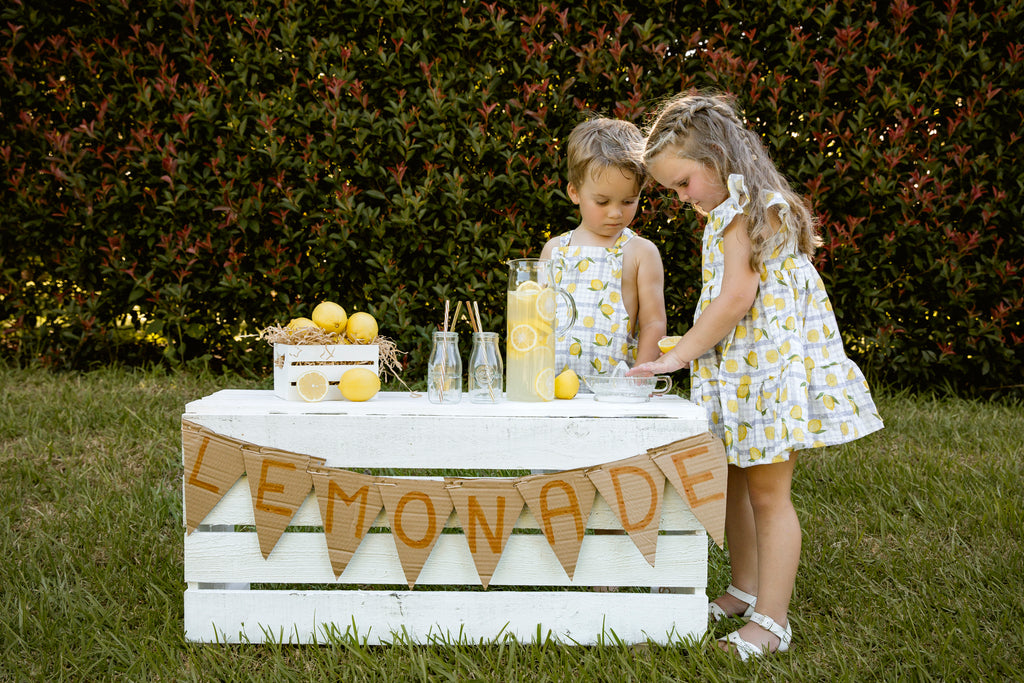 Lemonade print - a textured white linen base with a blue gingham and hand-drawn lemons. Handmade into a tiered skirt dress with frill cap sleeves. Handmade into short leg, rolled hem slouch cut overalls with tie top adjustable straps. Kids making lemonade at a roadside stand.