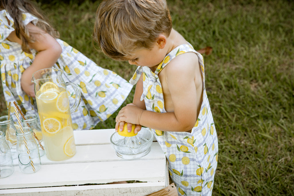 Lemonade print - a textured white linen base with a blue gingham and hand-drawn lemons. Handmade into short leg, rolled hem slouch cut overalls with tie top adjustable straps. Kids making lemonade at a roadside stand. Australian made. Boy making lemonade 