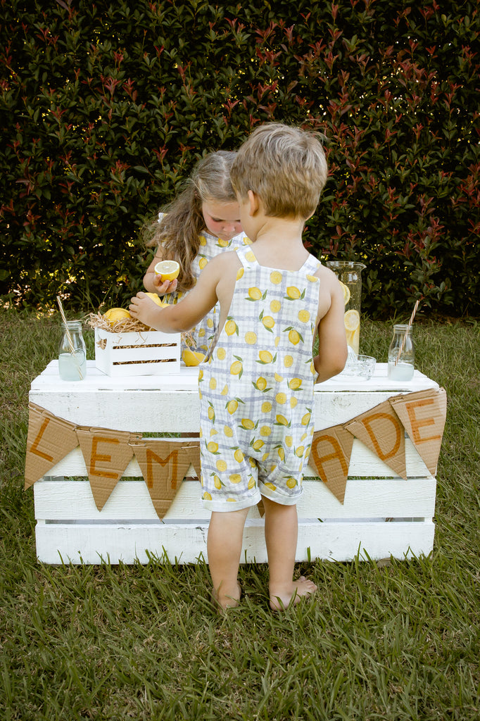 Lemonade print - a textured white linen base with a blue gingham and hand-drawn lemons. Handmade into short leg, rolled hem slouch cut overalls with tie top adjustable straps. Kids making lemonade at a roadside stand. Australian made.