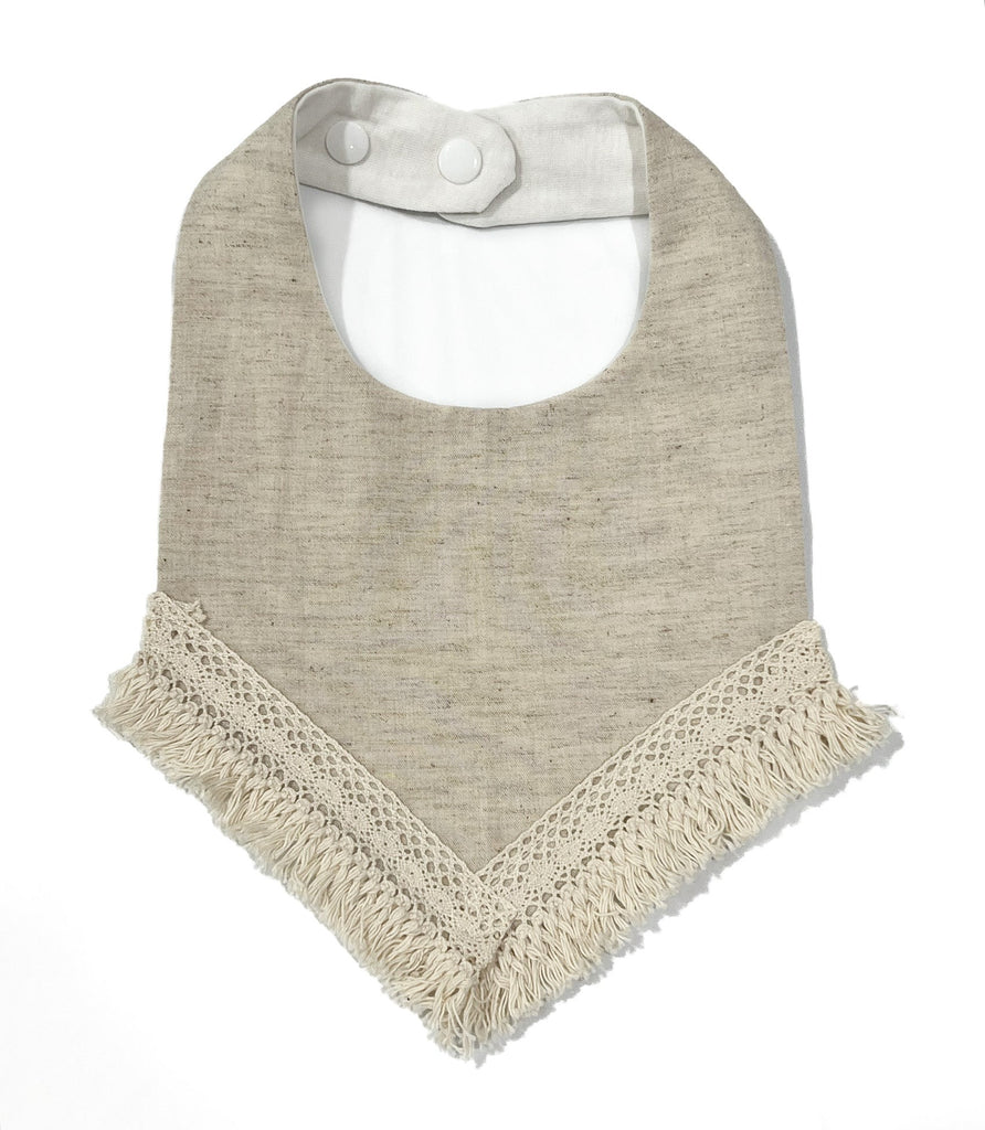 Oatmeal - natural, textured, neutral finish cotton . Handcrafted into a children’s bib with a tassel edge and adjustable clip back.