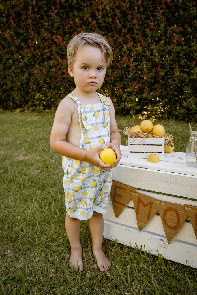 Lemonade print - a textured white linen base with a blue gingham and hand-drawn lemons. Handmade into short leg, rolled hem slouch cut overalls with tie top adjustable straps. Kids making lemonade at a roadside stand, boy holding lemon. Australian made.